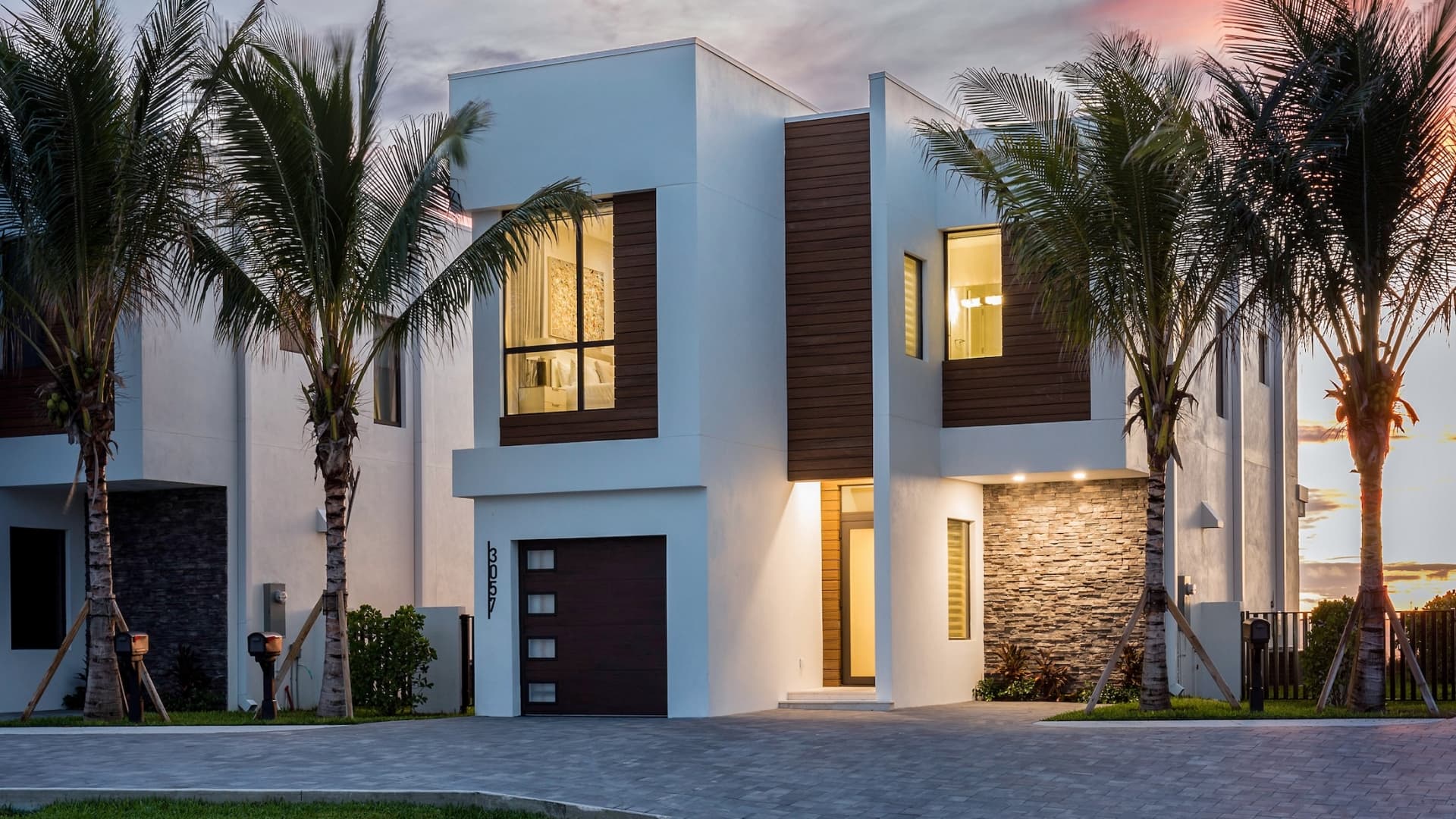 A multi-story luxury beach villa with an ultra-modern white and wood architectural design, a paved driveway, and palm trees lit up at dusk.