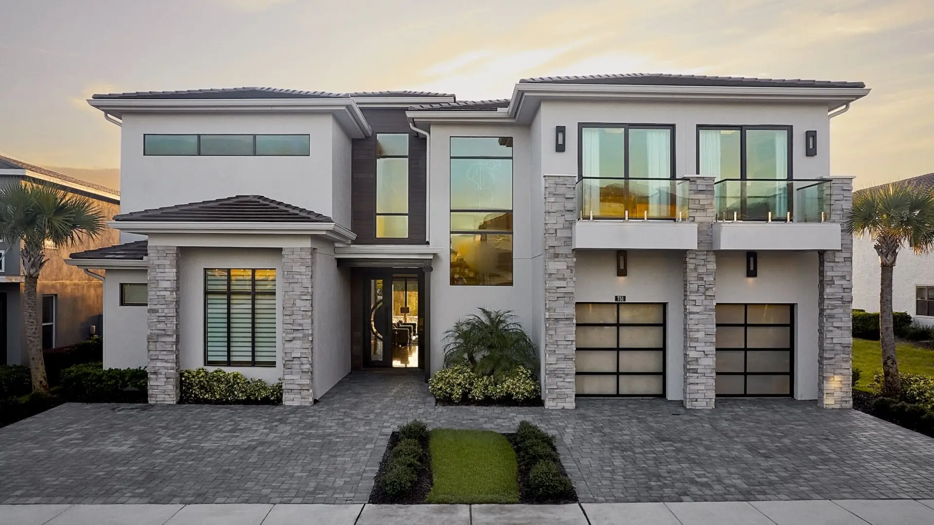 The street-front exterior of a grand, two-story luxury estate featuring a paved brick driveway, multiple stone-accent pillars, glass balconies, and two garages under a soft sunset sky.