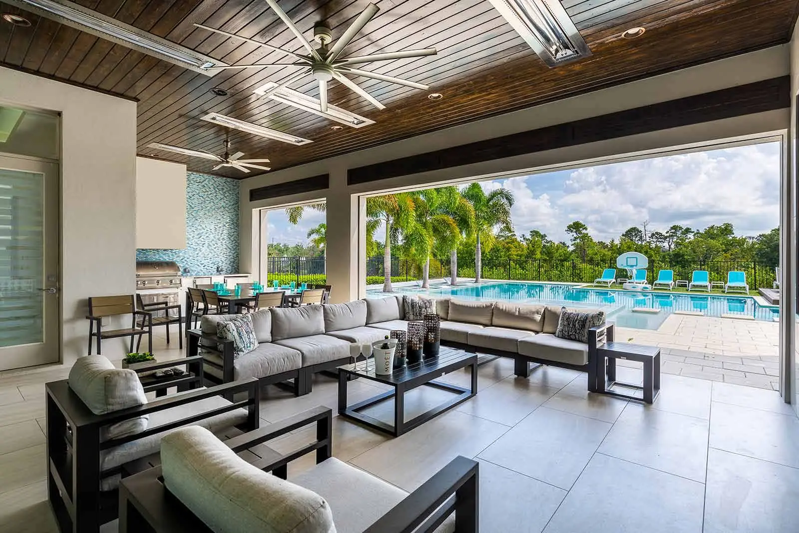 luxury covered patio with a dark wood ceiling, large sectional sofa, and outdoor kitchen, overlooking a private swimming pool with palm trees in the background.