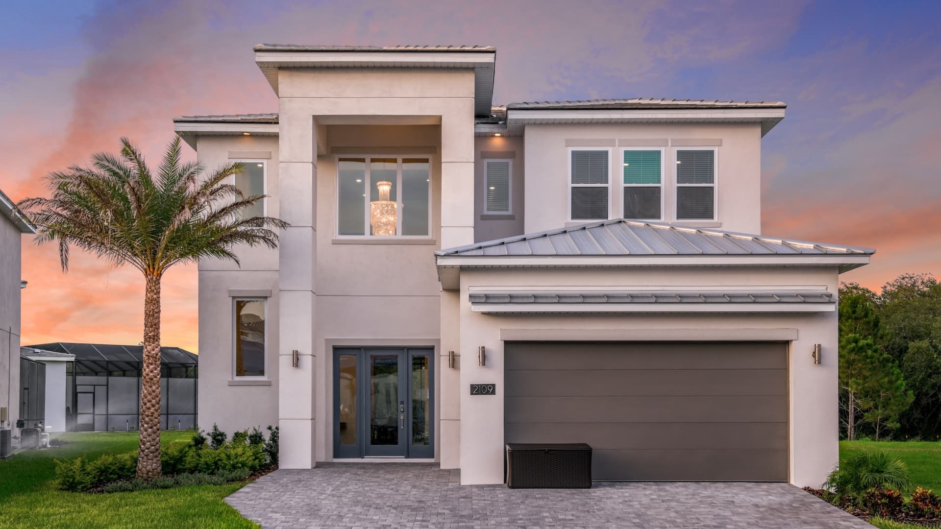A contemporary two-story resort home with a gray two-car garage, white stucco exterior, a small palm tree on the lawn, and a large statement chandelier glowing through the upper window at twilight.