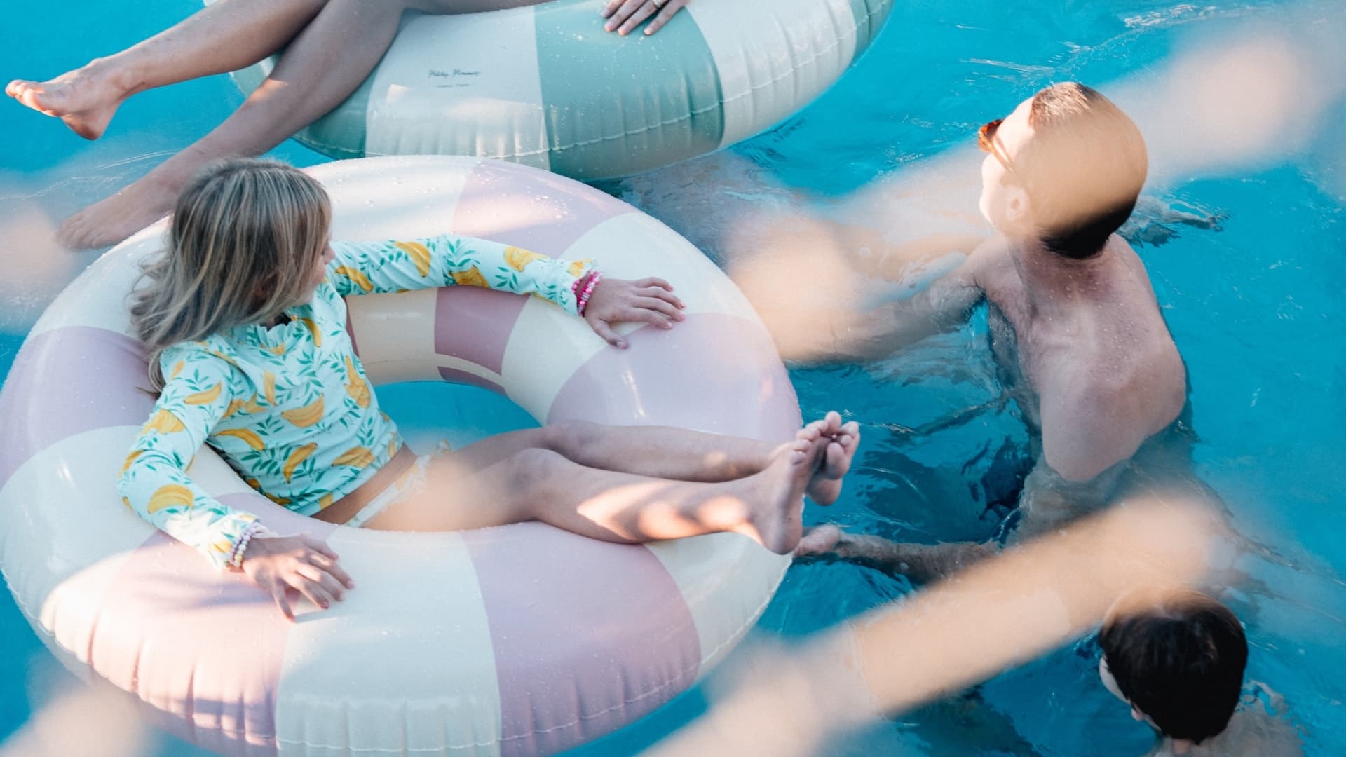 High-angle shot of a family of four enjoying a resort pool, with a woman and young girl floating on striped inner tubes while a man and boy swim nearby in the clear blue water.