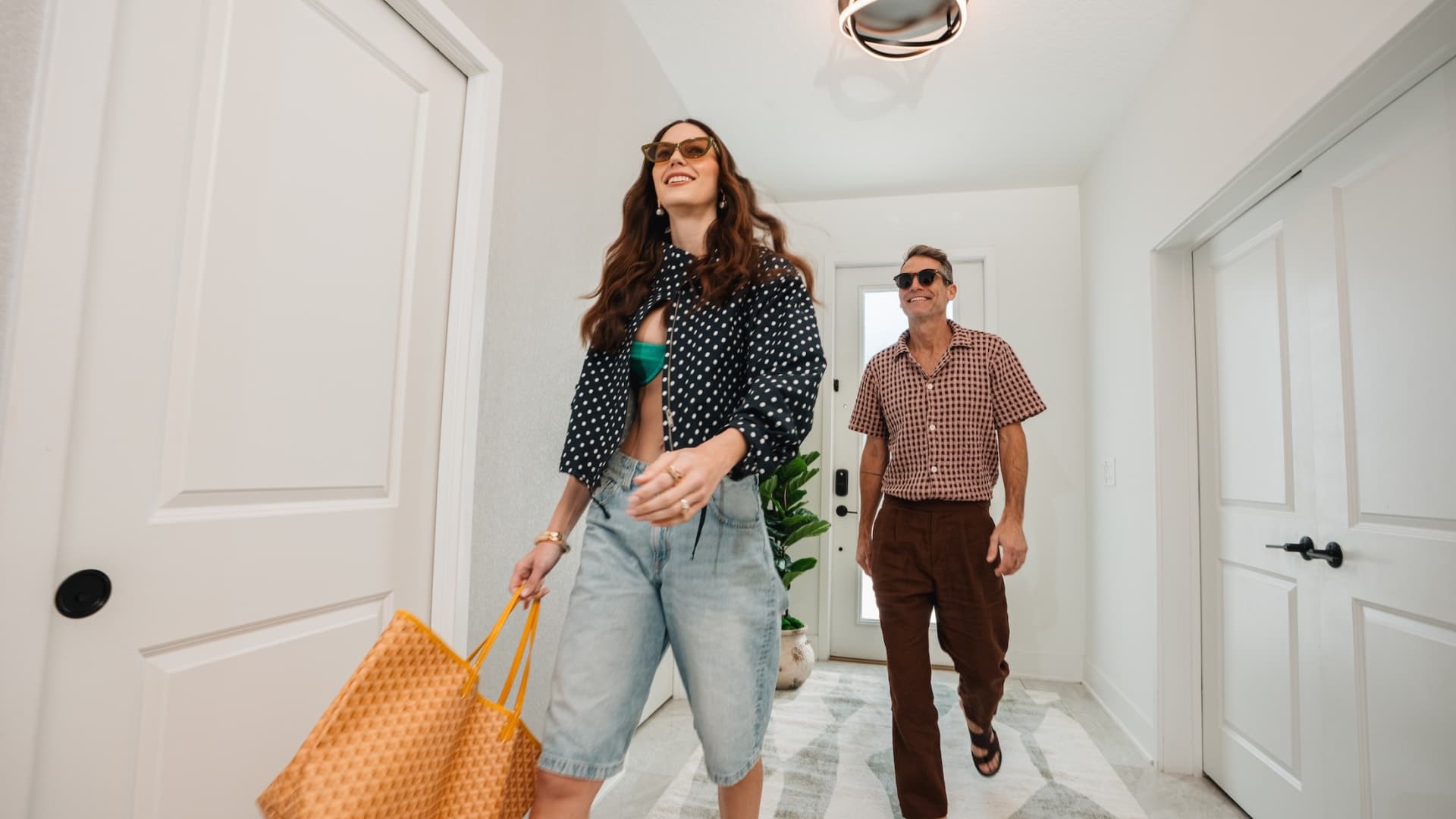 A stylish couple wearing sunglasses and summer attire walks through a bright, modern hallway of a vacation rental.