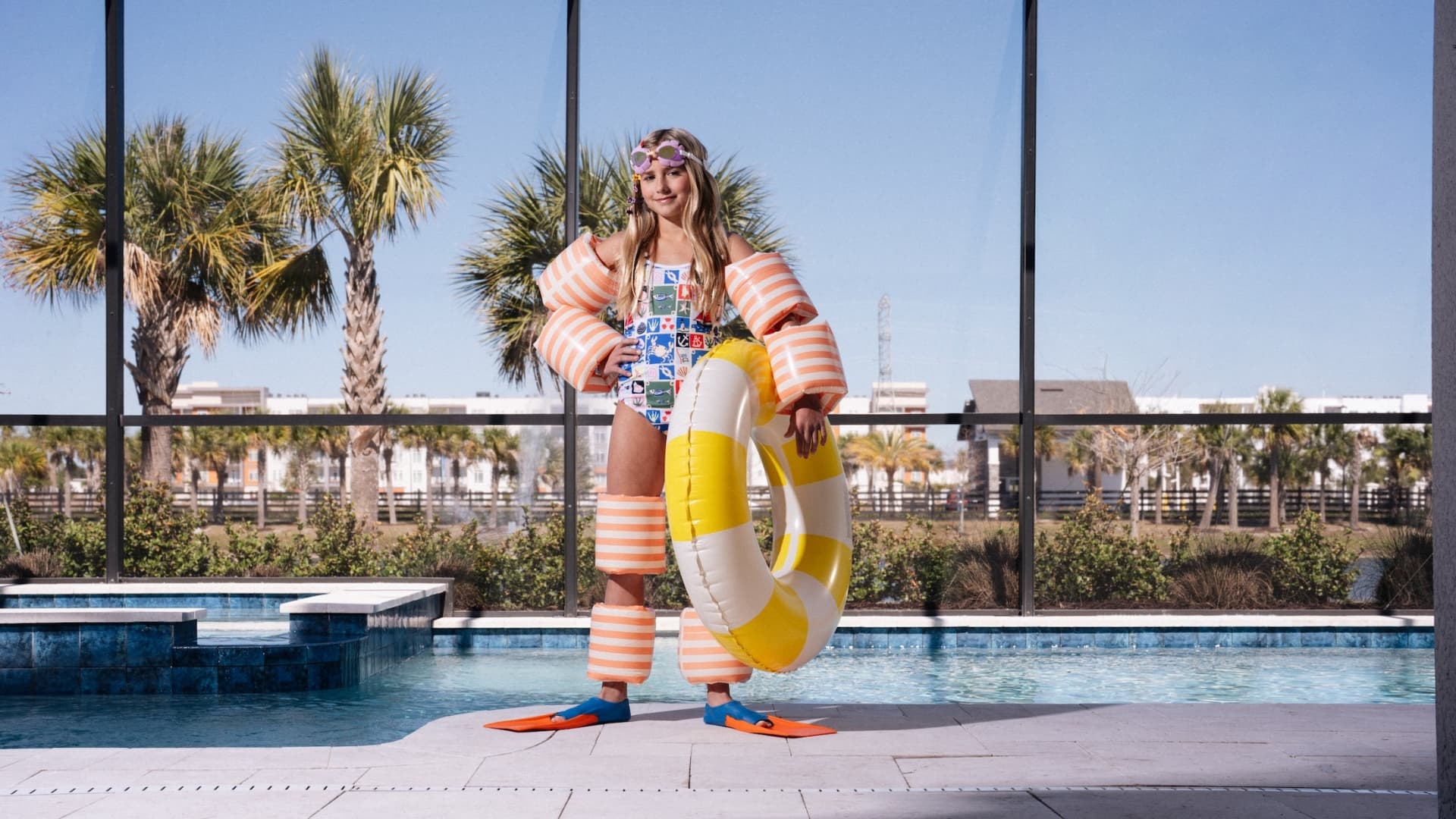 A young girl in a patterned swimsuit and orange flippers stands by a private screened-in pool holding a yellow inner tube.