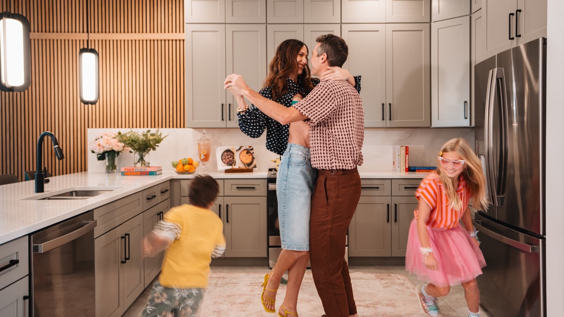 A family enjoying a vacation stay, with parents dancing in a modern kitchen while two young children play around them.