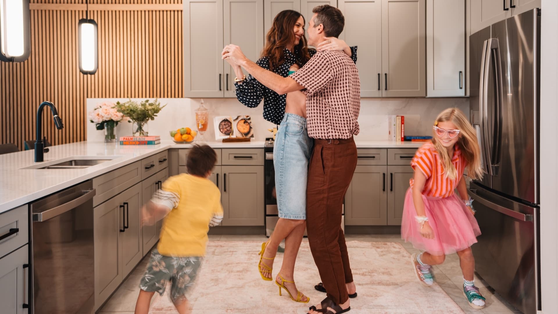 A family enjoying a vacation stay, with parents dancing in a modern kitchen while two young children play around them.